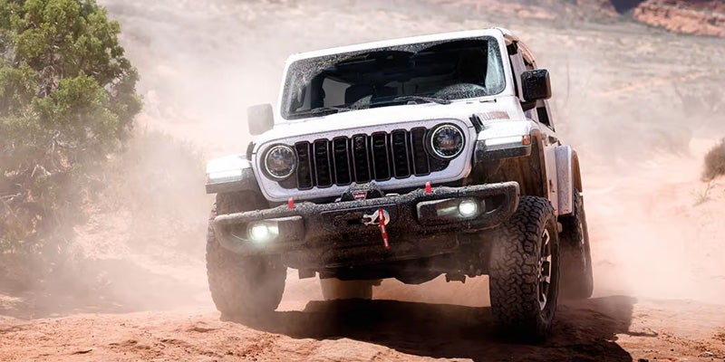 White Jeep Wrangler driving off-road through dusty desert terrain.